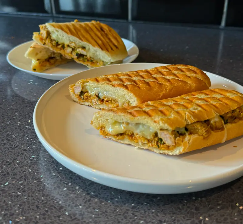 Two toasted panini on white plates, grill-marked bread cut open to show melted cheese and spiced chicken